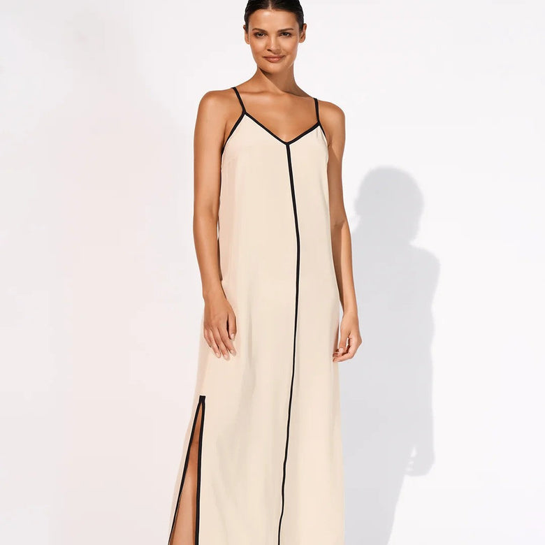 A woman modeling a long cream maxi dress with thin black trim and a high side slit, standing against a plain white studio background.