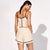 A woman stands barefoot against a white studio background, photographed from the back to show a light beige two-piece outfit with black trim. The set includes a spaghetti-strap camisole top and matching shorts.