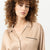 A woman with voluminous curly brown hair wearing a beige satin button-up shirt with black piping details, photographed against a clean white background.