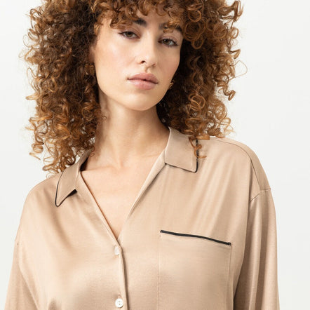 A woman with voluminous curly brown hair wearing a beige satin button-up shirt with black piping details, photographed against a clean white background.
