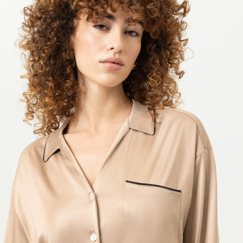 A woman with voluminous curly brown hair wearing a beige satin button-up shirt with black piping details, photographed against a clean white background.