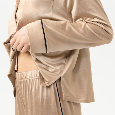 A close-up of a person wearing a beige satin pajama set with black piping trim, including a long-sleeve button-front top and matching pants.