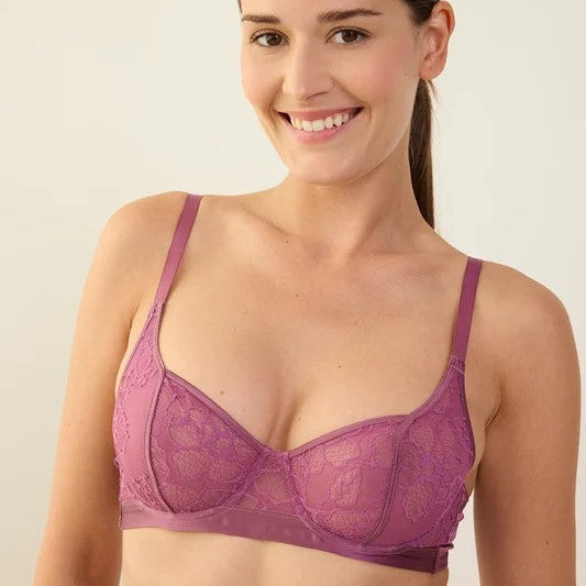 A smiling woman wearing a purple lace underwire bra against a plain light beige studio background.