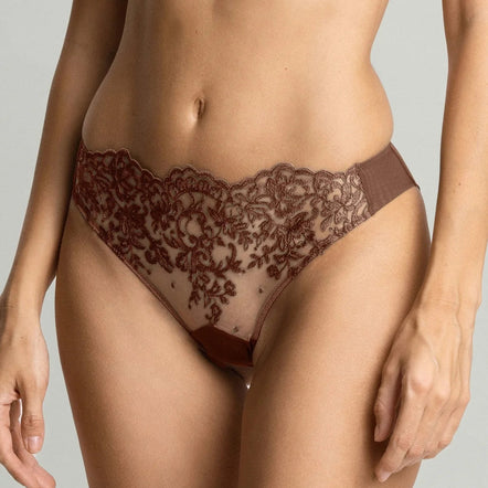 A close-up of a model wearing sheer brown lace underwear with a scalloped edge and a matching bra top, shown against a light gray studio background.
