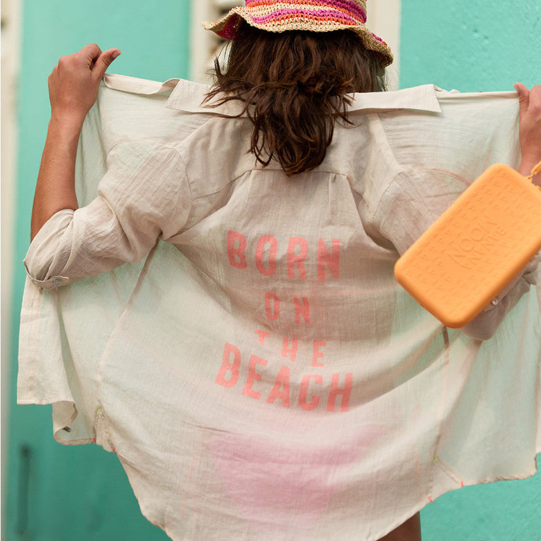 Person wearing a shirt with 'Born on the Beach' text, holding a pink bucket hat and orange item against a teal wall.
