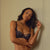 A woman modeling a black lace lingerie set consisting of a bra and matching thong, standing against a cream-colored paneled wall with a wall sconce visible on the right.