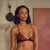 A woman modeling a burgundy lace lingerie set consisting of a triangle bra and matching panties, standing in a room with a fireplace and a large textured red painting on the wall.