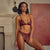 A woman modeling a burgundy lace lingerie set consisting of a triangle bra and matching panties, standing in a room with a fireplace and a large textured red painting on the wall.