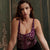 A woman wearing a purple and gold patterned corset top with black bottoms, leaning on a wooden table in front of an ornate green wall.