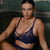 A woman sits on a dark green velvet armchair wearing a navy blue lace lingerie set with matching black pointed-toe heels, posed against a dark green paneled wall.