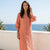 A woman wearing a long, peach-colored linen shirt dress with puff sleeves and a button-down front, standing on a stone path with a white wall and the blue ocean in the background.