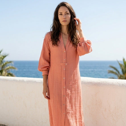 A woman wearing a long, peach-colored linen shirt dress with puff sleeves and a button-down front, standing on a stone path with a white wall and the blue ocean in the background.