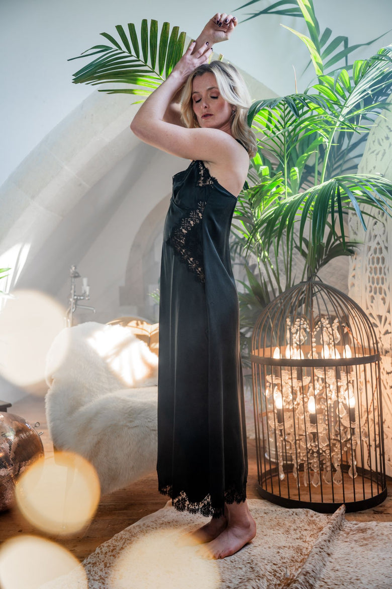 A woman wearing a black satin slip dress with lace trim stands in a styled room with palm leaves, a lit birdcage lamp, and a white chair in the background.
