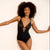 A woman with a large curly afro poses in a black lace-trimmed bodysuit with cutout details at the bust, paired with black knee-high boots, standing against a white wall with wooden parquet flooring.