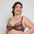 A plus-size woman modeling a matching brown sheer mesh and lace bra and panty set against a light gray studio background.