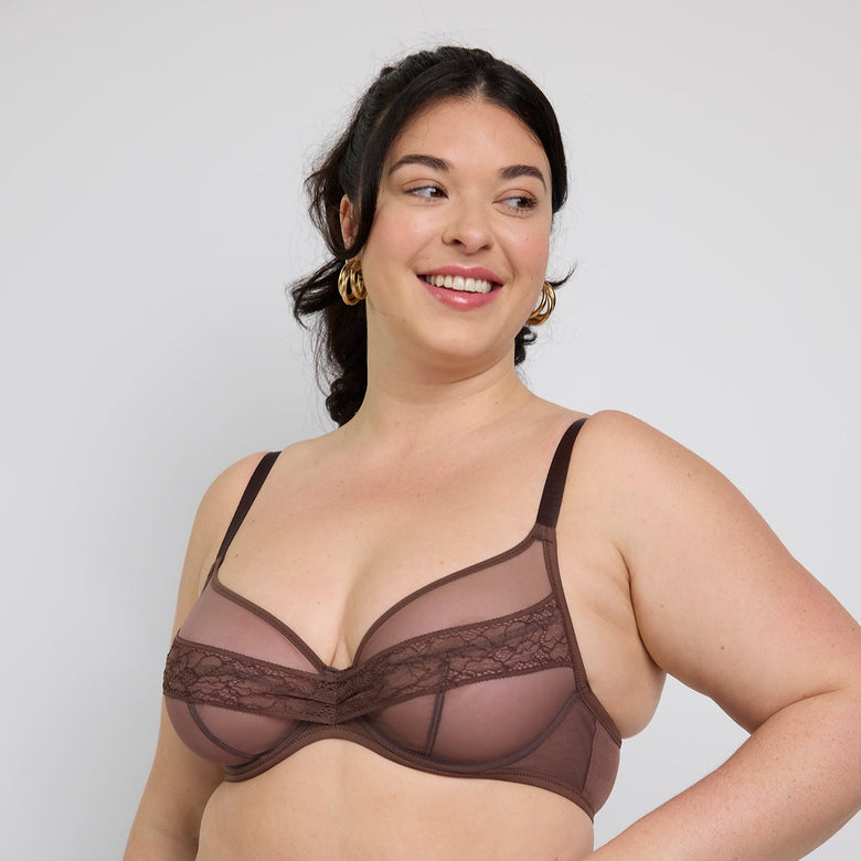 A plus-size woman modeling a matching brown sheer mesh and lace bra and panty set against a light gray studio background.