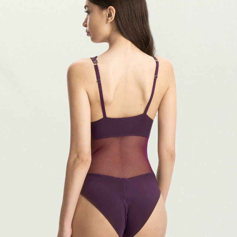 A woman shown from the back wearing a dark purple one-piece bodysuit with thin shoulder straps and a sheer mesh back panel, photographed against a plain light gray studio background.