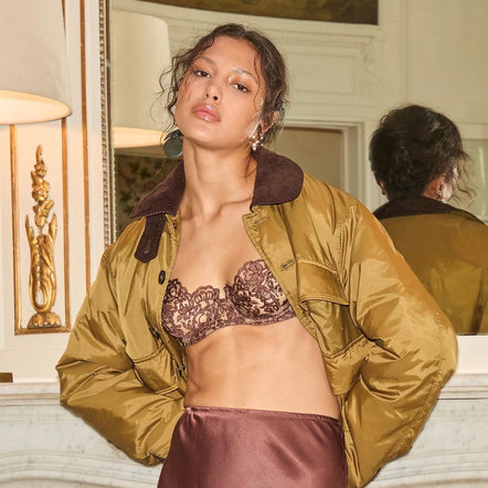 A model in an elegant room wearing a bronze lace bralette under an olive gold satin bomber jacket and a matching satin skirt.