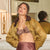 A model in an elegant room wearing a bronze lace bralette under an olive gold satin bomber jacket and a matching satin skirt.