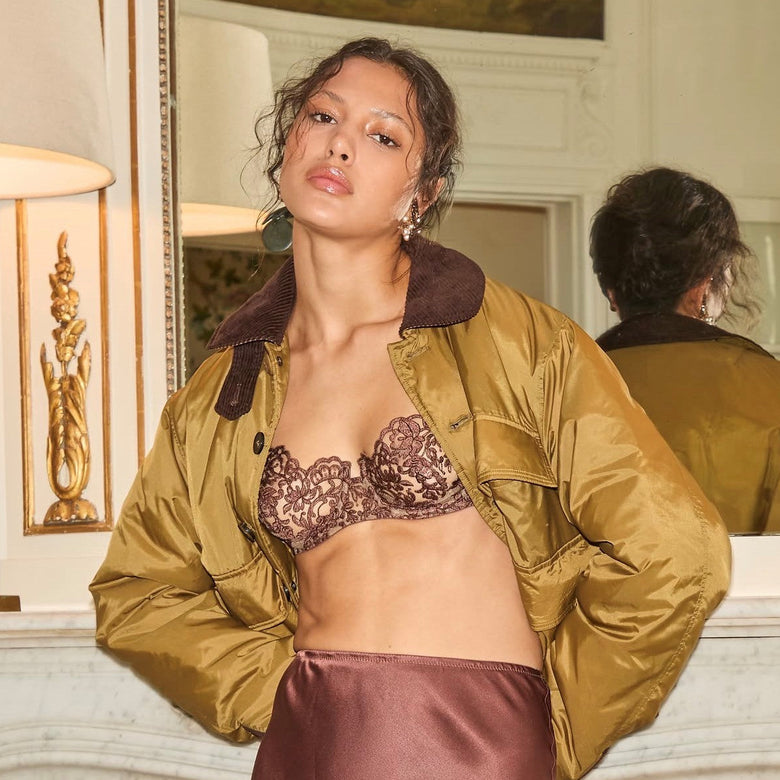A model in an elegant room wearing a bronze lace bralette under an olive gold satin bomber jacket and a matching satin skirt.