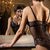 A woman wearing a black lace and mesh lingerie set is shown from behind, leaning on a gold-framed mirror in an ornate room. Her reflection is visible in the mirror, showing the front of the black lace bodysuit.