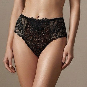 A woman stands barefoot in a studio wearing a black lace lingerie set consisting of a bra and matching high-waisted briefs against a plain beige wall and wood floor.