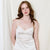 A woman with red hair stands against a cream fabric backdrop wearing a white satin camisole and matching lace-trimmed shorts.