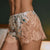 A close-up of a person wearing satin and lace sleep shorts. The shorts feature a floral print on a light cream satin base with prominent peach-beige floral lace panels and a scalloped lace hem.