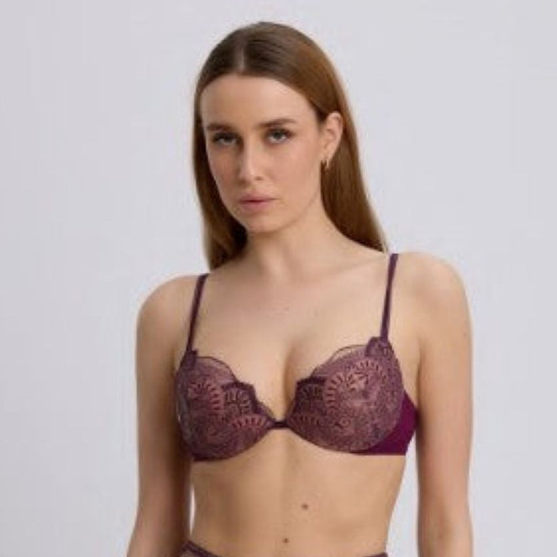 A woman modeling a matching lace lingerie set consisting of a sheer underwire bra and high-waisted briefs in a deep plum purple color, set against a plain light gray studio background.