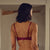 A woman stands with her back to the camera in a warmly lit living room, wearing a deep burgundy lace lingerie set with a strappy open-back bra and matching panties.