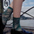 A person seated outdoors by a railing over water, wearing dark teal lace socks with a floral pattern and black loafers.