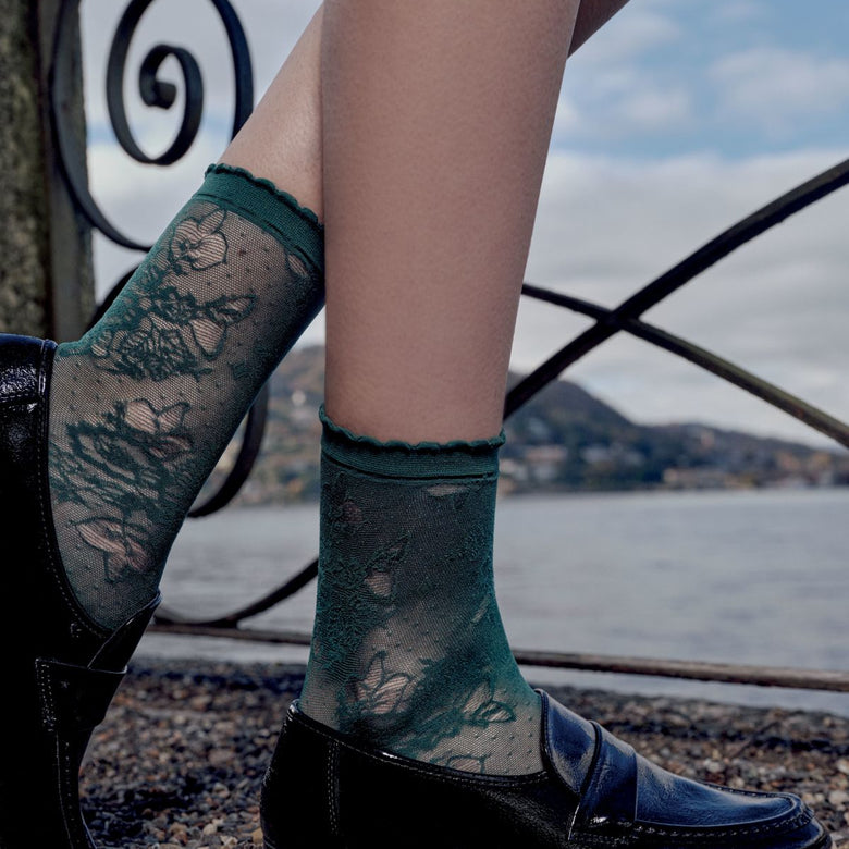 A person seated outdoors by a railing over water, wearing dark teal lace socks with a floral pattern and black loafers.