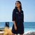 A woman wearing a navy blue short shirt dress with white stripe details on the chest stands outdoors near a pool and ocean, with a white lounge chair and dry grass in the background.