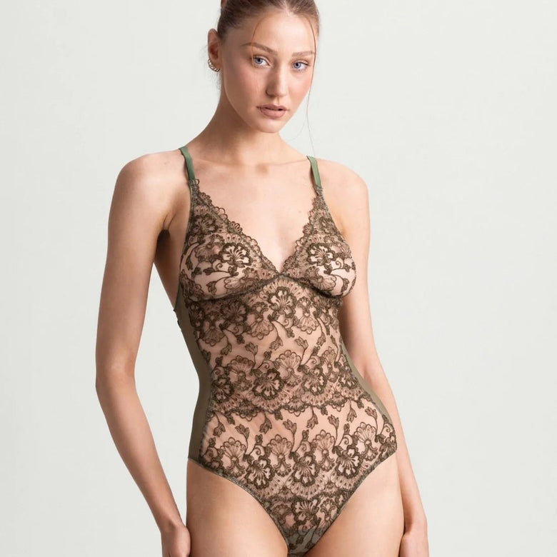 A woman modeling a sheer lace bodysuit in a warm taupe-brown color against a light gray studio background.