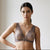 A woman modeling a sheer lace underwire bra in a muted mauve-purple shade, standing against a bright, minimalist off-white wall.