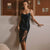A woman models a black satin slip dress with a sheer lace hem, standing in a minimalist room with three framed art pieces on the wall.