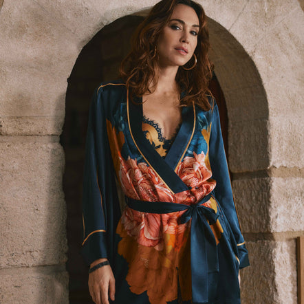 A woman models a short wrap-style romper with a dark teal blue base and a large orange, pink, and yellow floral print, featuring gold trim, posed in front of a rustic stone archway.