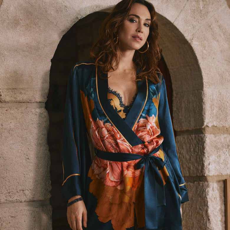 A woman models a short wrap-style romper with a dark teal blue base and a large orange, pink, and yellow floral print, featuring gold trim, posed in front of a rustic stone archway.
