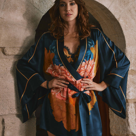 A woman wearing a dark blue satin robe with orange and pink floral print and gold trim, standing in front of a rustic stone archway.