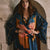 A woman wearing a dark blue satin robe with orange and pink floral print and gold trim, standing in front of a rustic stone archway.
