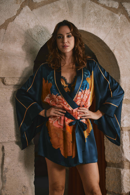 A woman wearing a dark blue satin robe with orange and pink floral print and gold trim, standing in front of a rustic stone archway.