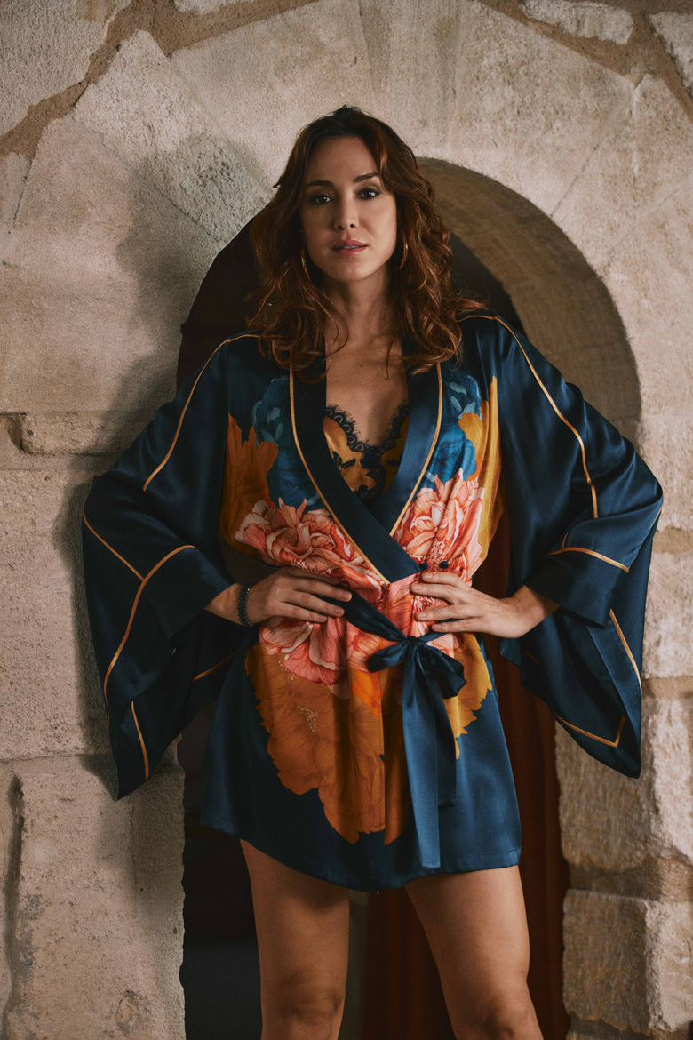A woman wearing a dark blue satin robe with orange and pink floral print and gold trim, standing in front of a rustic stone archway.