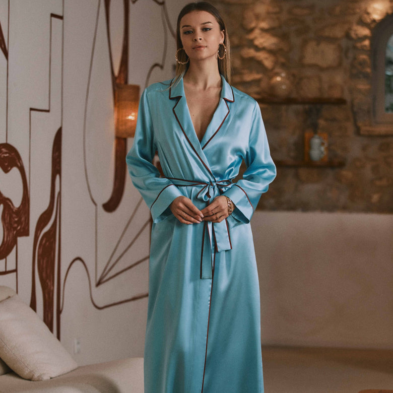 A woman wearing a light blue satin robe with dark piping and a matching tie belt, standing in a modern living room with a beige sofa, wooden table, and stone wall in the background.