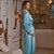 A woman wearing a light blue satin robe with dark piping stands in profile in a modern living room setting with a stone wall and abstract wall art.