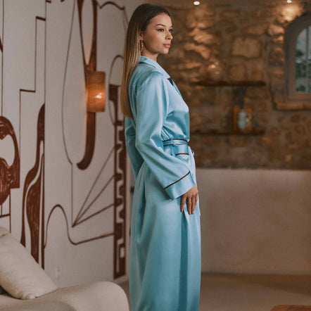 A woman wearing a light blue satin robe with dark piping stands in profile in a modern living room setting with a stone wall and abstract wall art.