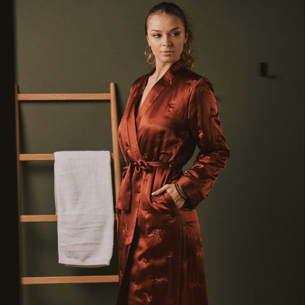 A woman wearing a long, satin-like rust-brown robe with a self-tie belt and subtle tonal floral pattern, standing in a dimly lit room with a wooden towel ladder and white towel in the background.