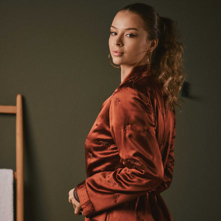 A woman wearing a rust-brown satin robe with tonal floral embroidery, standing in profile against a dark olive-green wall.