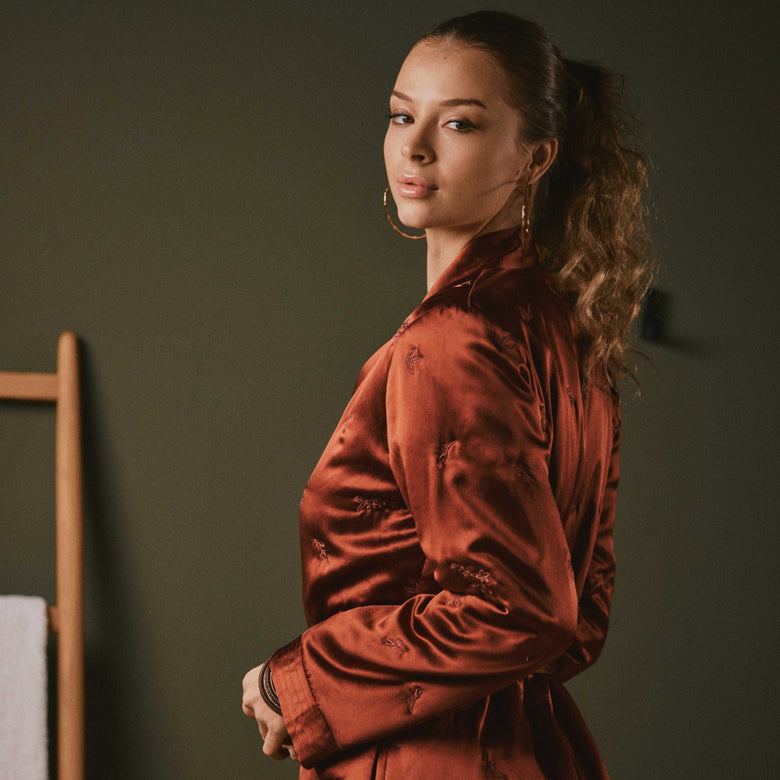 A woman wearing a rust-brown satin robe with tonal floral embroidery, standing in profile against a dark olive-green wall.
