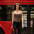 A woman walking on a city street in front of a red bus, wearing a strapless leopard-print corset top, black ripped skinny jeans, black ankle boots, and carrying a black tote bag.