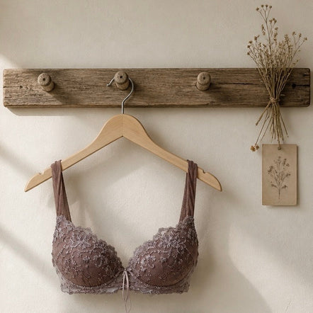 A lace underwire bra in a muted mauve-brown color hangs on a wooden hanger from a rustic wooden wall hook. The background is a warm off-white wall with a small dried floral decoration on the right.